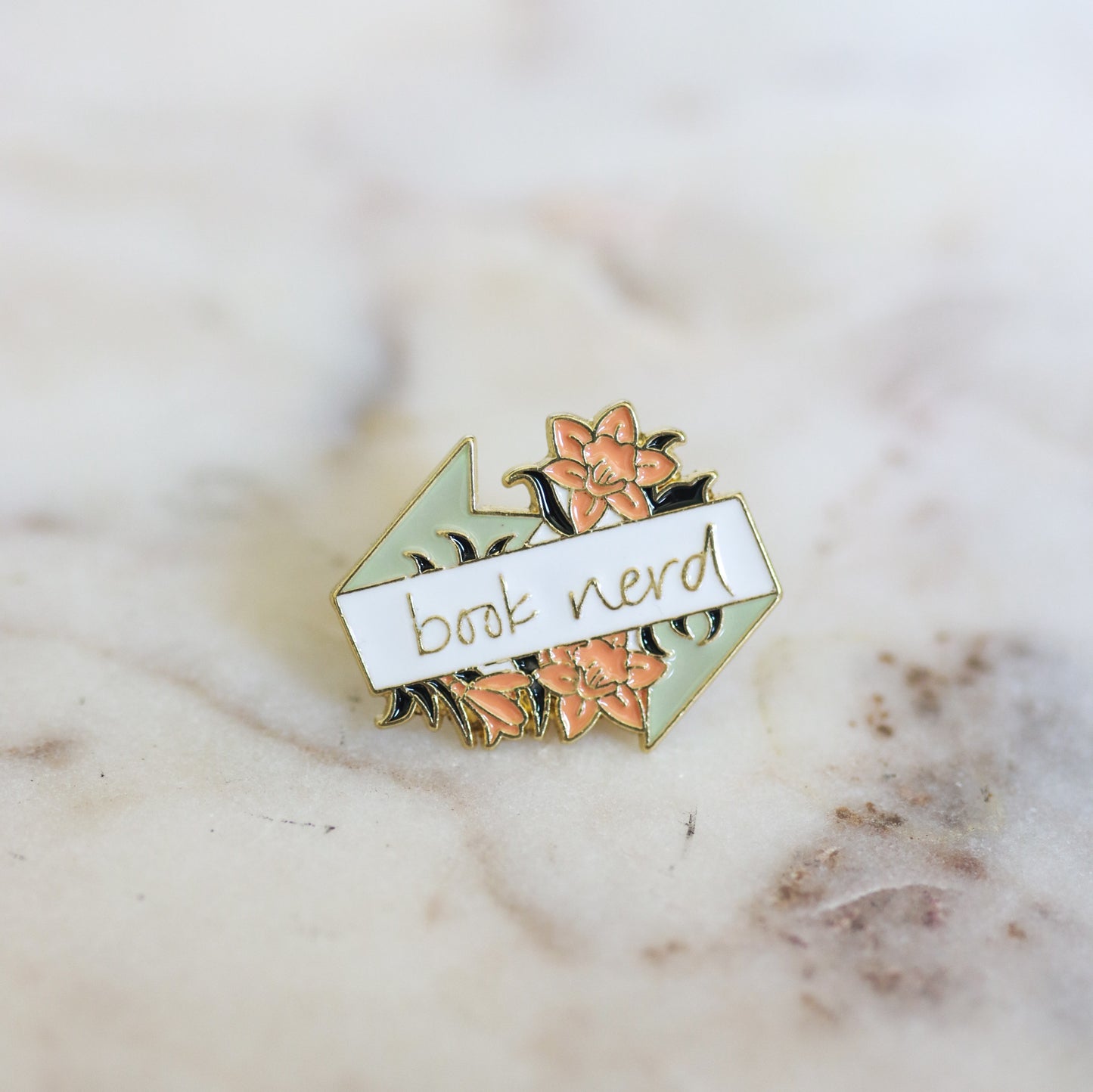 A pin shaped like flowers with a banner that reads Book Nerd in pink, mint, and white. From Copper Crow Swag Market's Book Pins collection
