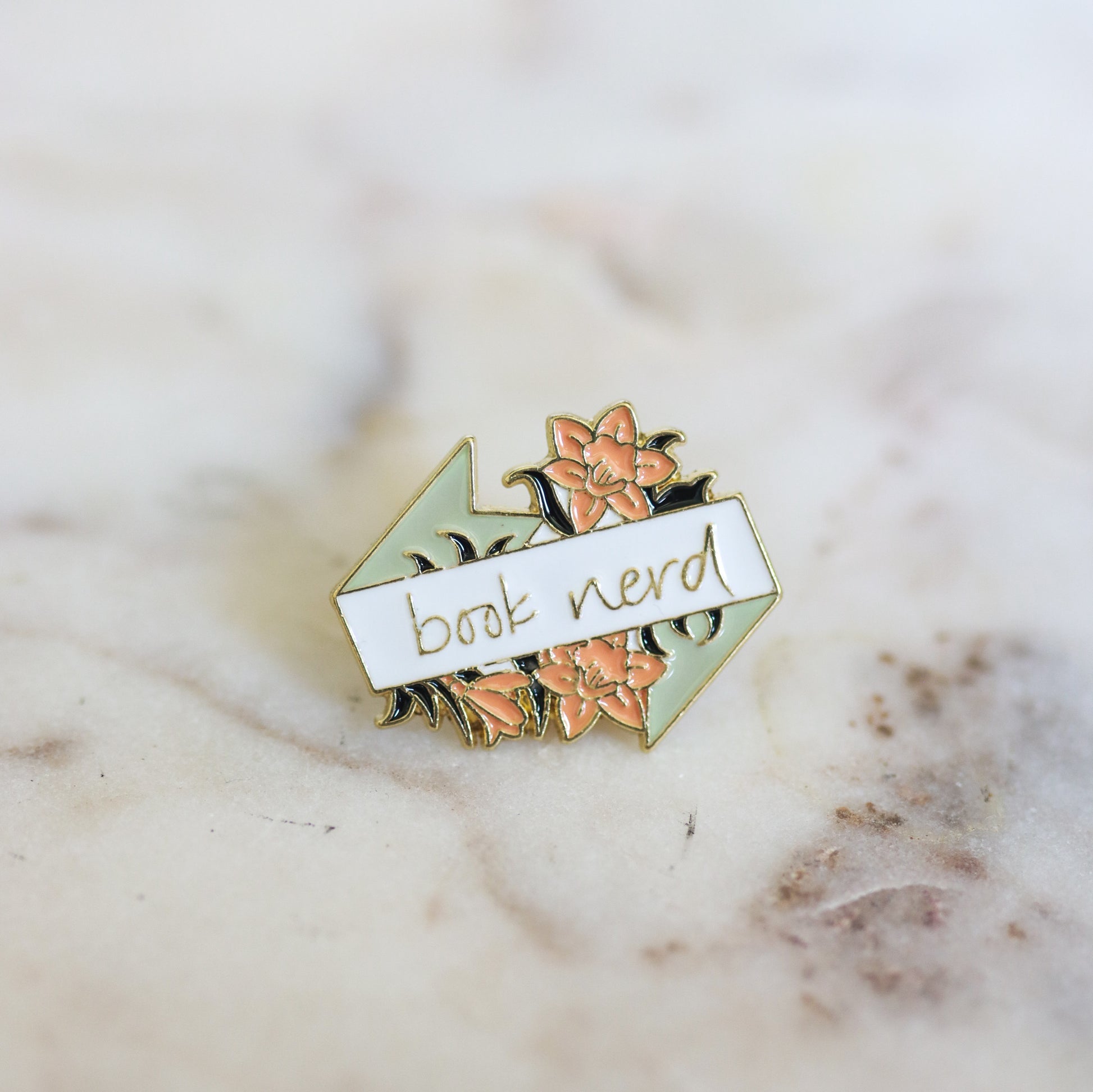 A pin shaped like flowers with a banner that reads Book Nerd in pink, mint, and white. From Copper Crow Swag Market's Book Pins collection