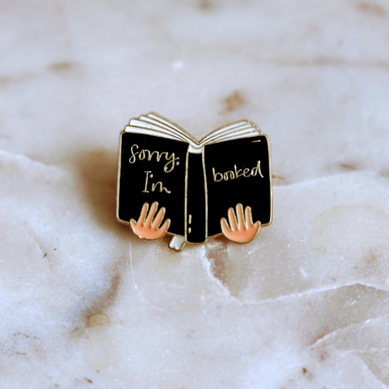 A pin shaped like hands holding a black open book with the phrase Sorry, I'm Booked. From Copper Crow Swag Market's Book Pins collection