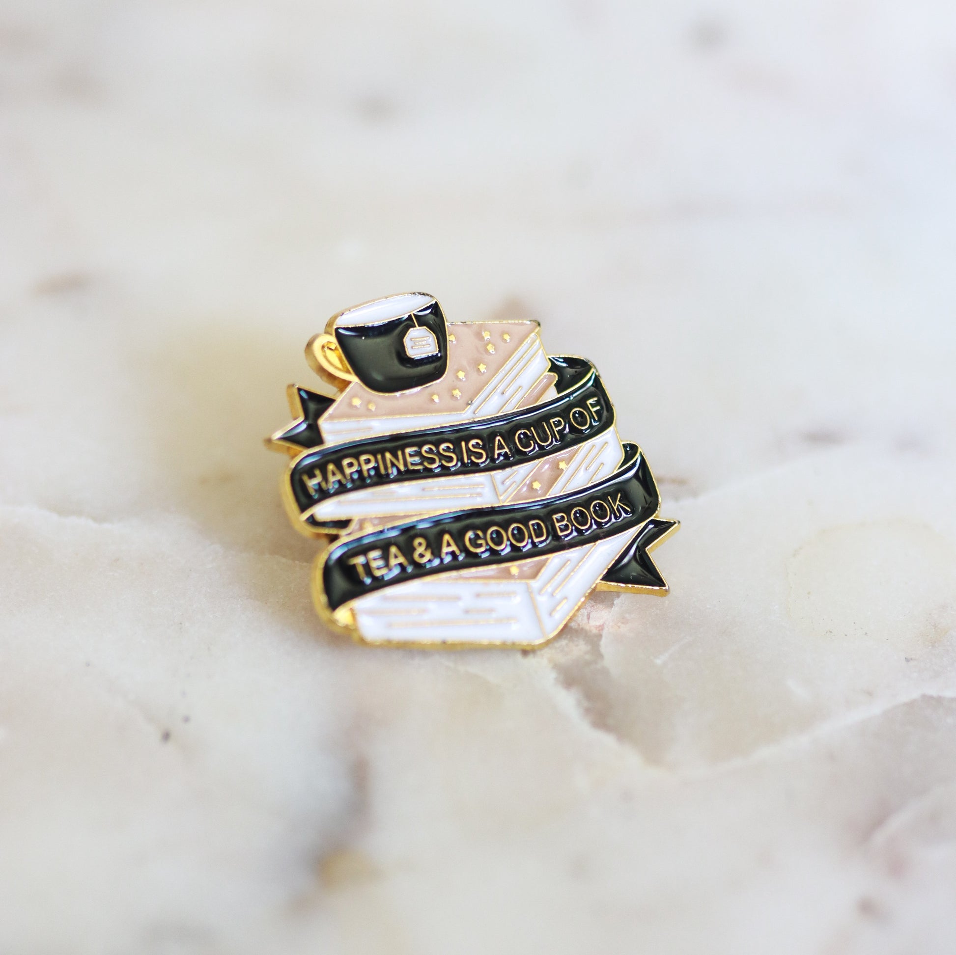 A pin shaped like a pink stack of books with a cup of tea and the phrase Happiness is a Cup of Tea and a Good Book. From Copper Crow Swag Market's Book Pins collection